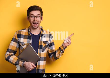 Der verblüffte junge Mann in einer Brille lächelte glücklich, hielt den Laptop und zeigte mit dem Zeigefinger auf freie freie freie freie Werbeträger für Werbetexte. WOW, OMG, unglaublich, Promotionkonzept Stockfoto