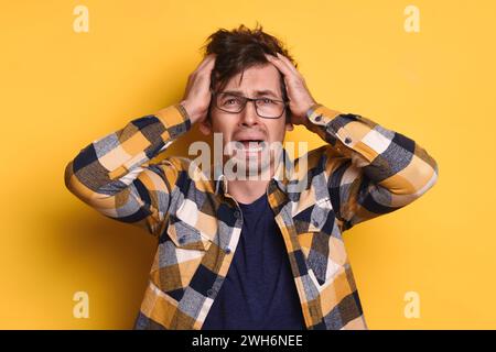 Frustrierter emotionaler Mann in Brille mit Händen auf dem Kopf, der sein Haar rauszieht, vor Verzweiflung, Trauer weint, nervös, Panik, er verlor sein Geld, bekam Ärger, isoliert an der gelben Wand Stockfoto