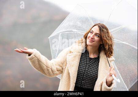 Eine fröhliche junge Frau hält einen transparenten Regenschirm und spürt die Regentropfen auf ihrer Hand. Stockfoto