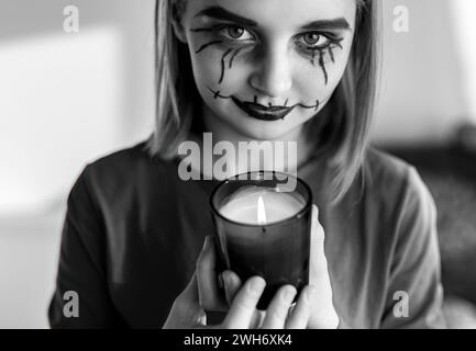 Ein junges Mädchen mit gruseligem Halloween Make-up posiert vor der Kamera mit einer Kerze. Kinderportrait für gruselige Ferien in Schwarz-weiß. Stockfoto