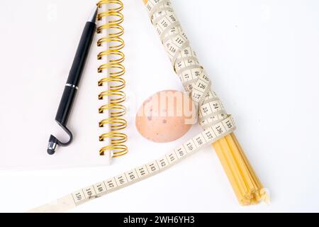 Ein Haufen roher Spaghetti mit Maßband, Hühnerei und Notizbuch zum Schreiben mit schwarzem Stift auf weißem Hintergrund Stockfoto