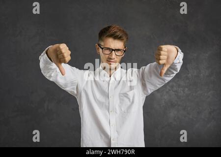 Enttäuschter junger Mann, der die Daumen nach unten zeigt und isoliert auf grauem Hintergrund steht Stockfoto