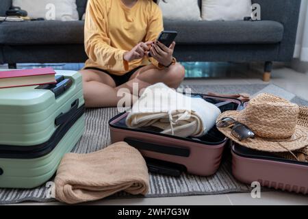 Junge asiatische Frau packt ihren Koffer für Sommerreisen und überprüft die Hotelreservierung auf dem Handy Stockfoto