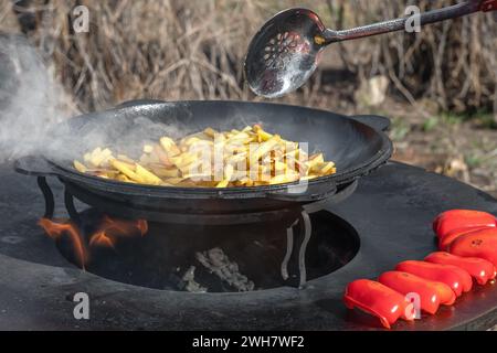 Gemüse, Paprika und Kartoffeln werden an einem runden Kamin im Freien gebraten. Runder Grill in Form einer Schüssel mit Feuer im Inneren. Rundes Metallholz Stockfoto