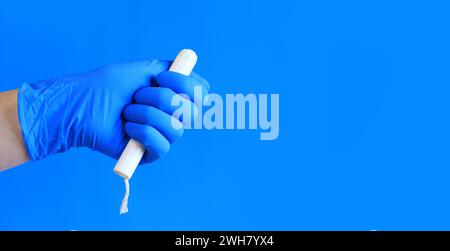 Tampon in einem Pappappappapplikator in der Hand einer Frau, blauer Hintergrund. Eine Hand in einem blauen medizinischen Latexhandschuh hält einen Tampon. Weibliche Körperhygiene PR Stockfoto