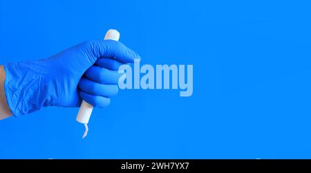 Tampon in einem Pappappappapplikator in der Hand einer Frau, blauer Hintergrund. Eine Hand in einem blauen medizinischen Latexhandschuh hält einen Tampon. Weibliche Körperhygiene PR Stockfoto