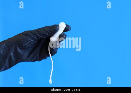 Weibliche Hand mit einem Tampon-Nahaufnahme auf blauem Hintergrund. Eine Hand in einem medizinischen Handschuh aus schwarzem Latex hält einen Tampon. Weibliches Körperpflegeprodukt Stockfoto