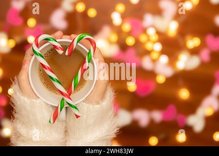 Frauenhand mit Pullover, der eine Kaffeetasse mit Zuckerstangen in Herzform hält, defokussiertes Bokeh-Licht für Valentinstag-Feier dekoriert Stockfoto