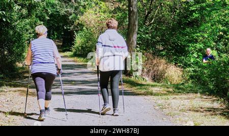 Ein gruseliger Mann versteckt sich im Wald, während zwei Damen mit nordic Walking-Stöcken auf einem Waldweg am Rheinufer in Deutschland vorbeigehen. Stockfoto