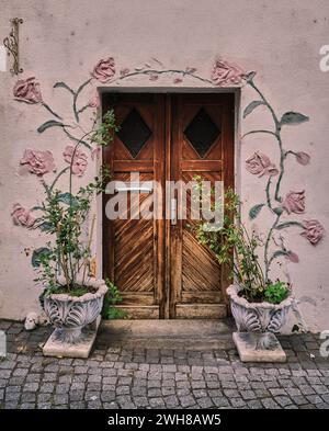 Charaktervolle Doppeltür aus Holz mit zwei kunstvollen Blumentöpfen mit Blumen und kunstvoller Verzierung von Rosenreben auf der pinkfarbenen Stuckfassungstür. Stockfoto