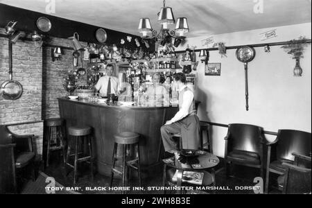 Dorset. c.1960 – Eine echte Fotopostkarte mit dem Titel „Toby Bar, Black Horse Inn, Sturminster Marshall, Dorset“, die die „Toby Bar“ im Black Horse Inn zeigt, einem alten kutschenhaus in Sturminster Marshall an der A350 zwischen Poole und Blandford Forum. Die Bar ist mit einer Reihe von Krügen und Tankstellen ausgestattet, darunter ein paar toby-Krüge. Die Besitzer John und Jean Sibley stehen hinter der Bar. Stockfoto