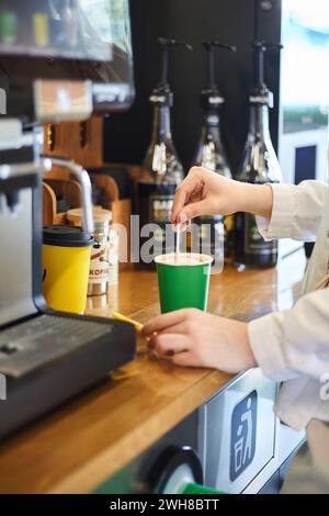 Hände der jungen Frau, die Kaffee macht, Rührzucker umrühren. Einwegbecher aus grünem Papier. Selbstbedienungsstelle für heiße Getränke mit Kaffeemaschine und Sirupe. Café, Stockfoto
