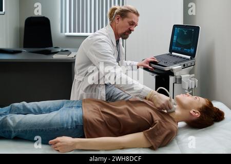 Reife Endokrinologin im Laborkitt bewegte Ultraschallsonde am Hals einer jungen Patientin im Sitzen am Ultraschallgerät Stockfoto