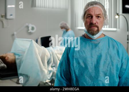 Schwerer männlicher Chirurg in medizinischen Peelings und Schutzkappe, die Kamera gegen Operationstisch mit Serviette überzogen Stockfoto