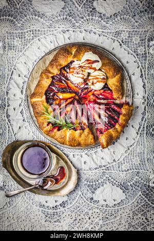 Rustikale Plum Galette, in Scheiben geschnitten, mit Vanilleeis, Rotwein-Karamellsauce und einem frischen Rosmarinzweig auf kompliziertem Spitzenhintergrund Stockfoto