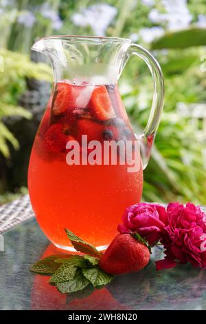 Beeren- und Rosenblütenspritzer, erfrischendes Sommergetränk. Kännchen in der Nähe im Garten im Freien. Stockfoto