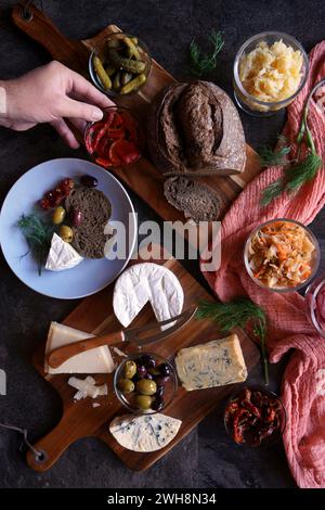 Gesunde probiotische Käseplatte serviert mit Kimchi, Sauerteigbrot mit dunkler Färbung und fermentiertem Gemüse. Männliche Hand, die nach Nahrung greift. Stockfoto