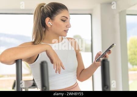 Junge Frau mit sportlichem Top, die Musik auf ihrem Handy wählt, um sie mit ihren kabellosen Kopfhörern im Fitnessstudio während des Trainings zu hören Stockfoto