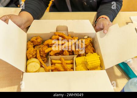 Hände einer nicht erkennbaren Person, die eine Fastfood-Box öffnet, mit Huhn, Mais und Pommes frites auf einem Tisch Stockfoto