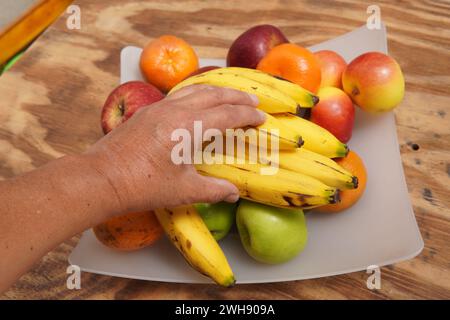 Die Hand des Mannes pflückt Bananen in einer Obstschale mit Äpfeln und Mandarinen Stockfoto