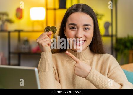 Glückliche Kaukasierin, freischaffende Entwicklerprogrammiererin, die zwei Goldmünzen hält. Mädchen-Aktienhändler, die Bitcoins verdienen, nachdem sie den Handel online überwacht haben, sitzen am Tisch im Home Office Stockfoto