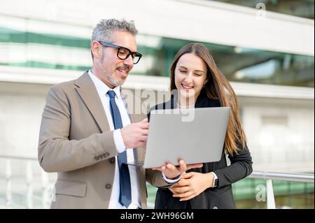 Ein leitender Geschäftsmann und eine jüngere Kollegin beschäftigen sich gemeinsam mit Inhalten auf einem Laptop im Freien und repräsentieren Mentoring und Zusammenarbeit in einer modernen Geschäftsumgebung Stockfoto