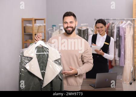 Reinigungsservice. Glücklicher Mann, der drinnen einen Kleiderbügel mit Jacke hält. Mitarbeiter macht sich Notizen am Arbeitsplatz Stockfoto