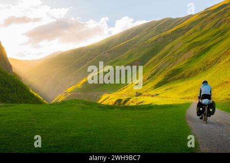 Radfahrer Rückblick auf der Straße in der malerischen kaukasus Natur mit malerischem Sonnenuntergang Hintergrund im Tal. Reisende auf dem Fahrrad. Alleinreisefahrrad für Langstrecken Stockfoto