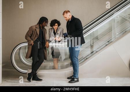 Drei Geschäftsleute in stilvoller Winterkleidung, die auf einer Rolltreppe zu einem zwanglosen Geschäftstreffen stehen, arbeiten fokussiert und unkompliziert zusammen. Stockfoto
