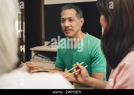 Ein japanischer Mann, der zu Hause mit Essstäbchen isst, schaut und lächelt seine Familie an. Stockfoto