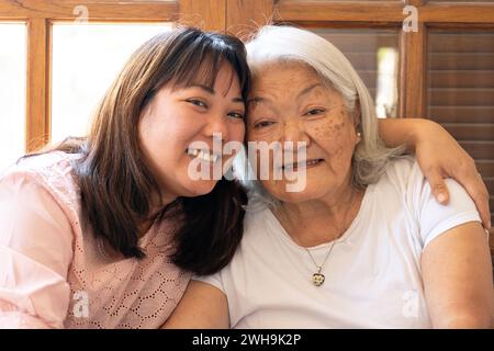 Nahaufnahme einer japanischen erwachsenen Tochter, die ihre grauhaarige Mutter umarmt, lächelt und in die Kamera blickt. Stockfoto