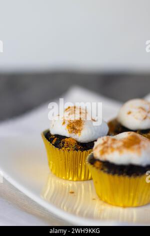 Schokoladen-Cupcakes mit weißer Marshmallow-Glasur, echtes essbares Blattgold streut in einer goldenen Cupcake-Verpackung auf einer weißen Platte mit Platz für Text Stockfoto