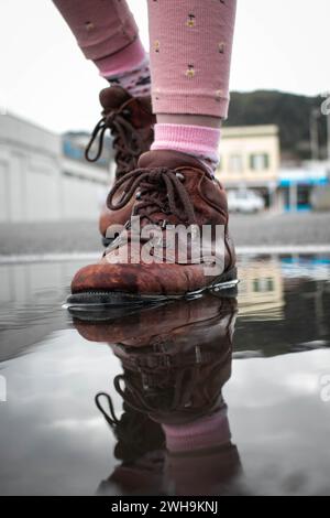 Nahaufnahme eines Mädchens in rosa Strumpfhosen mit nassen braunen Stiefeln, das sich in einer Regenpfütze auf der Straße spiegelt. Stockfoto