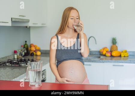 Eine schwangere Frau, die die lebenswichtigen Vorteile des Wassers während der Schwangerschaft berücksichtigt, steht mit einem Glas in der Küche und unterstreicht die entscheidende Rolle der Hydratation bei Stockfoto