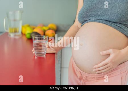 Eine schwangere Frau, die die lebenswichtigen Vorteile des Wassers während der Schwangerschaft berücksichtigt, steht mit einem Glas in der Küche und unterstreicht die entscheidende Rolle der Hydratation bei Stockfoto