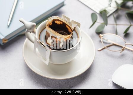 Eine Glasschale frisch gebrühten Kaffees mit einem praktischen Tropfkaffefilter auf dem Bürotisch. Kaffeepause, schnelle Art, heißes Getränk zuzubereiten Stockfoto
