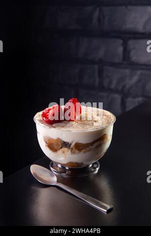 Tiramisu-Dessert in einer Glasschale, dekoriert mit Kaffee und Erdbeeren auf schwarzem Tisch mit Backsteinwand dahinter Stockfoto