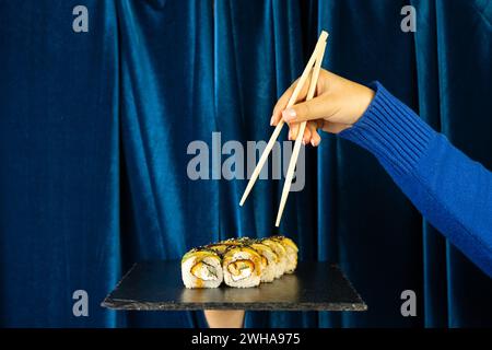 Weibliche Hand mit Sushi und Holzstäbchen auf tiefblauem Hintergrund mit Vorhang. Asiatisches Restaurant-Menü-Konzept Stockfoto