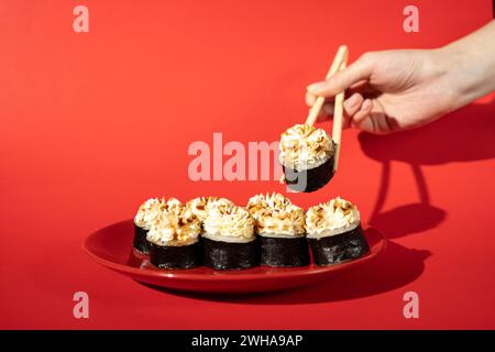 Weibliche Hand, die gebackene Sushi-Rolle mit Stäbchen auf rotem Hintergrund mit hartem Schatten hält Stockfoto