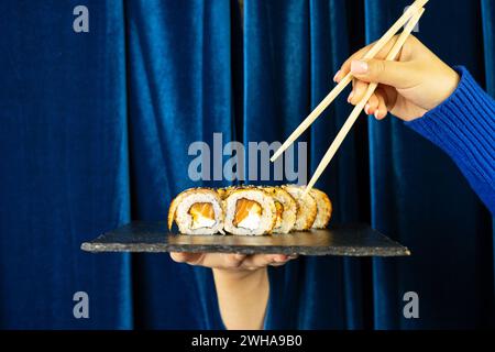 Weibliche Hand mit Sushi und Holzstäbchen auf tiefblauem Hintergrund mit Vorhang. Asiatisches Restaurant-Menü-Konzept Stockfoto