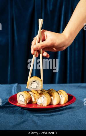 Die weibliche Hand hält Philadelphia Sushi-Brötchen mit geräuchertem Aal, Avocado und Frischkäse mit Bambusstäbchen auf blauem Samtgrund. Japanisches Essen. Stockfoto