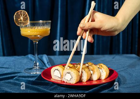Sushi auf einem roten Teller auf blauem Samtgrund, weibliche Hand nimmt Sushi-Rolle mit Essstäbchen, Nahaufnahme Stockfoto