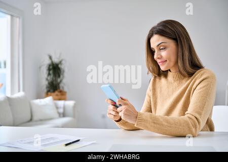 Glückliche reife Frau, die ihr Handy benutzt, und im Wohnzimmer zu Hause am Tisch sitzt. Stockfoto