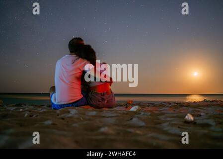 Ein romantisches Paar umarmt sich von hinten auf dem mondbeleuchteten Sand am Meer und schafft eine intime Verbindung unter dem leuchtenden Leuchten des Vollmondes. Stockfoto