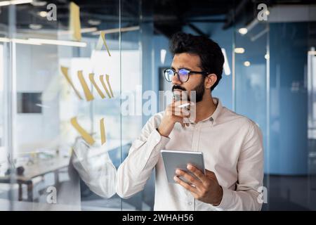 Nachdenklicher und konzentrierter junger Indianer, der im Büro neben Glasplatte mit Tablett steht und Notizen ansieht. Denkt über neue Ideen und Problemlösungen nach. Stockfoto