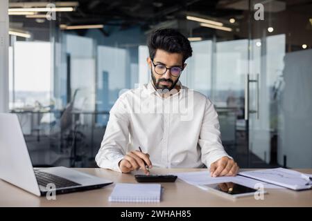 Ein indischer Geschäftsmann mit Bart arbeitet intensiv an seinem Laptop, umgeben von einem modernen Büroinnenraum, das für engagierte Professionalität steht. Stockfoto