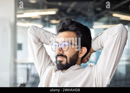 Selbstbewusster indischer Geschäftsmann mit Bart, der sich an seinem Schreibtisch in einer modernen Büroumgebung entspannt, Hände hinter dem Kopf machen eine Pause. Stockfoto