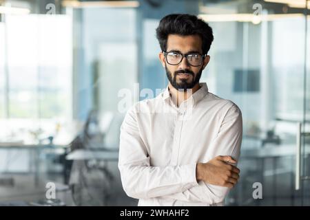 Ein Angestellter, der ein Hemd trägt, mit Bart und Brille, steht in einem Büro, die Arme überkreuzt und ein Lächeln trägt. Stockfoto