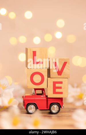 Rotes Truck-Spielzeug mit Holzwürfel und Text Love auf der Rückseite des Pickups, um Liebe zum Valentinstag mit unscharfem weißem Blumenfeld und d zu liefern Stockfoto