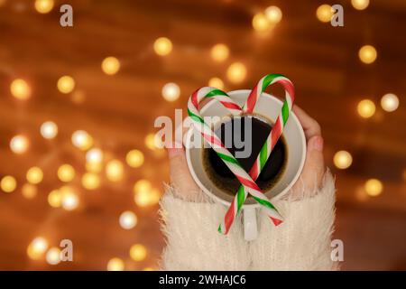 Frauenhand mit Pullover, der eine Kaffeetasse mit Zuckerstangen in Herzform hält, defokussiertes Bokeh-Licht für Valentinstag-Feier dekoriert Stockfoto